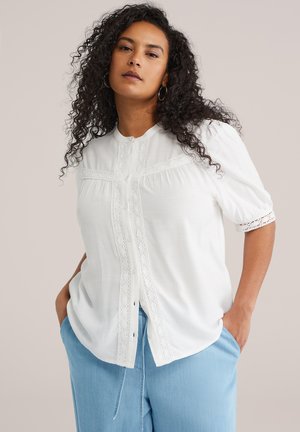 Kobieta z długimi kręconymi włosami, ubrana w białą, zapinaną na guziki bluzkę z koronkowymi detalami oraz jasnoniebieskie spodnie, ręce w kieszeniach, neutralne tło.