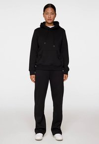 Czarna bluza z kapturem i kieszenią z przodu, z sznurkiem w kapturze, zestawiona z pasującymi czarnymi spodniami. Oba elementy wykonane z miękkiego materiału. Białe trampki.