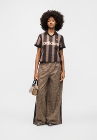 Brown and black striped short-sleeve polo shirt with "adidas" logo. Loose leopard print pants with side stripes. Model holds small handbag.