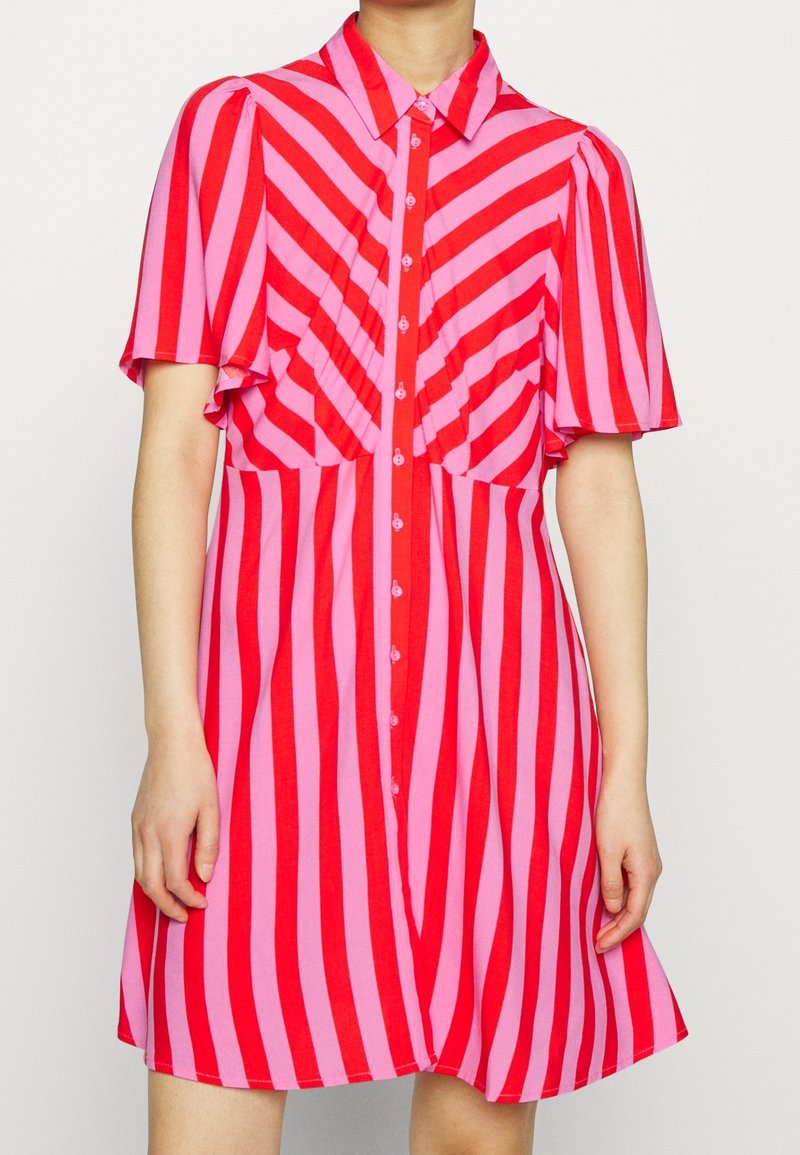 Robe à manches courtes avec des rayures diagonales et verticales rouges et roses, boutonnée devant, avec un col pointu, portée par une personne.
