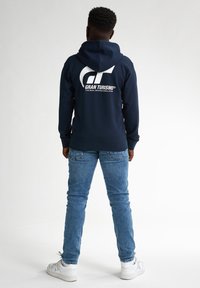 Felpa navy con grande logo bianco "Gran Turismo" sul retro. Abbinata a jeans blu e sneakers bianche, presenta un look casual.