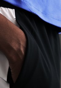 Χέρι που ακουμπάει μέσα στην τσέπη μαύρου παντελονιού, με ένα ανοιχτό μπλε πουκάμισο ορατό πάνω από τη μέση.