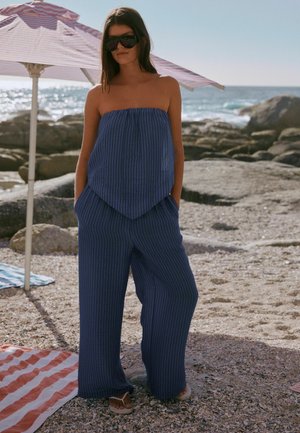 Woman wearing blue pinstriped strapless top and wide-legged pants, dark sunglasses, standing on rocky beach near striped umbrella and sea.