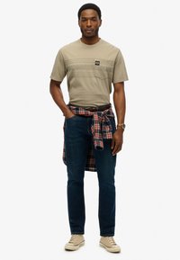 T-shirt di cotone beige con taschino sul petto, jeans blu scuro e camicia flanella a quadri rossa legata in vita. Sneakers beige completano l'outfit.