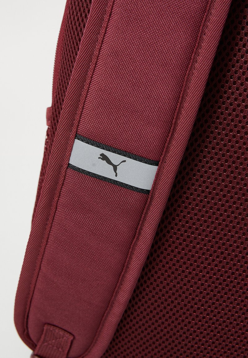 Roter Mesh-Rucksackgurt mit einer glatten, kastanienbraunen Stoffoberlage. Verfügt über ein graues Akzentband mit einem schwarzen Logo. Strukturierter Mesh für Atmungsaktivität.