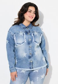 Chaqueta de mezclilla azul con patrones de estrellas, bordes deshilachados y acentos de strass. Dos bolsillos en el pecho con cierre de botón y un cuello tipo solapa.