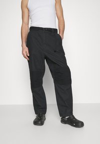 Czarne spodnie cargo o luźnym kroju, z łatami na kolanach, szlufkami na pasek oraz teksturowanym materiałem. Połączone z czarnymi klapkami.