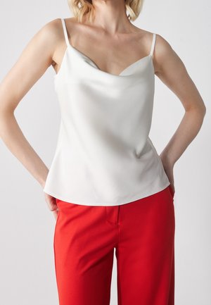 Mujer que lleva una blusa de satén blanca sin mangas con tirantes finos y pantalones rojos de talle alto, de pie con las manos en los bolsillos.