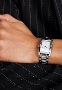 Silver rectangular watch with a textured silver band, featuring a white face, black hour markers, and a few decorative rhinestones.