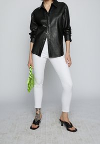 Camisa de cuero negra con botones, jeans skinny blancos, sandalias de tacón negras y un bolso verde con un diseño estampado y detalles de flecos.