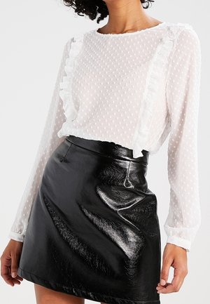 Femme portant un chemisier blanc texturé avec des détails à volants, associé à une jupe patineuse en cuir noir brillant.