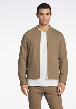 Veste zippée marron en tissu doux, avec un col rond et des poches latérales, associée à un t-shirt blanc et un pantalon assorti.