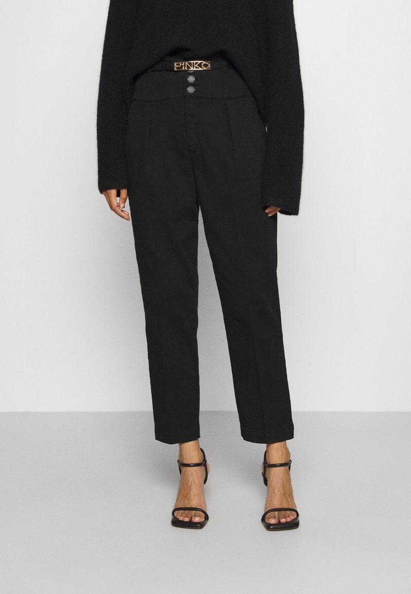 Femme portant un pantalon noir taille haute avec deux boutons et une ceinture à boucle dorée, associé à un haut noir à manches longues et des talons ouverts.