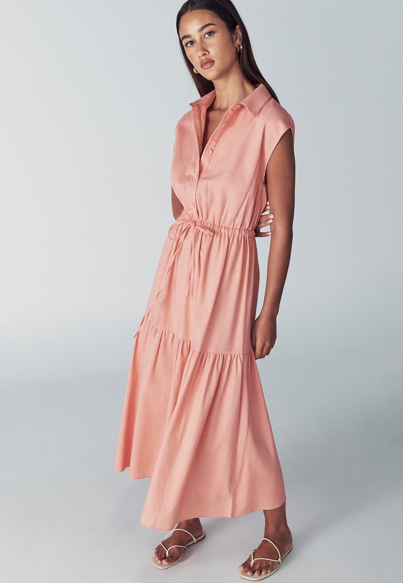 Różowa, warstwowa maxi sukienka z krótkimi rękawami, kołnierzykiem i regulowanym w pasie. Wykonana z lekkiego materiału, posiada detale z guzikami oraz zwiewną spódnicę.