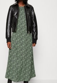 Schwarze Lederjacke in cropped Länge über einem grün gemusterten Maxikleid mit gewellten Linien, ergänzt durch eine silberne Kettenhalskette.