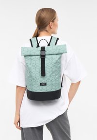 Mujer con cabello castaño claro recogido, vestida con una camisa blanca y pantalones grises, llevando una mochila enrollable de color verde claro y negro con estampado.