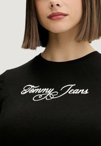 T-shirt nera a coste con logo "Tommy Jeans" in corsivo bianco sul davanti. Design aderente con scollo rotondo.