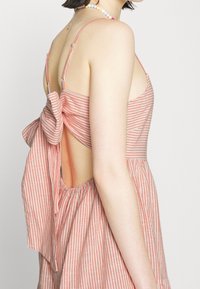 Raidallinen mekko vaalean korallin ja valkoisen puuvillan yhdistelmässä, jossa on ohuet olkaimet, nauha-akcentti selässä ja avonainen selkäosa.