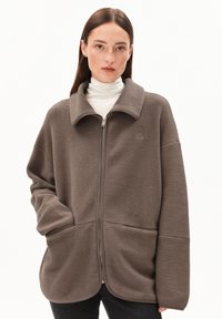 Femme portant une grande veste en polaire marron à fermeture éclair, avec un col haut et des poches, associée à un pull à col roulé blanc et un pantalon foncé, sur fond blanc.