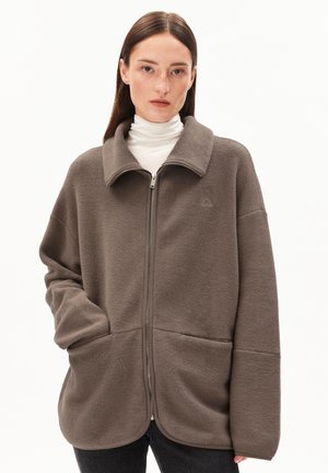 Vrouw in een oversized bruine fleecetrist met hoge kraag en zakken, gecombineerd met een witte coltrui en donkere broeken, tegen een witte achtergrond.