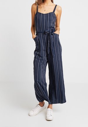 Femme portant une combinaison sans manches à rayures verticales bleu marine et blanches avec une ceinture nouée à la taille et des baskets blanches, debout les mains dans les poches.