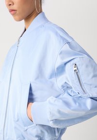 Jasnoniebieska kurtka bomberska wykonana z błyszczącego materiału. Posiada ściągacze na mankietach i kołnierzu, boczne kieszenie oraz kieszeń na rękawie na zamek.