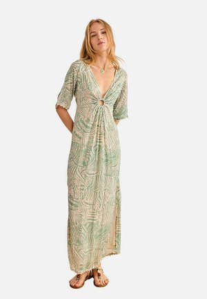 Mujer que lleva un vestido largo con un patrón en verde claro y beige, mangas hasta el codo, detalle de anillo en el frontal, abertura lateral y sandalias planas.