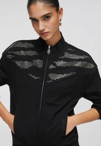 Veste noire à col montant, fermeture éclair, ornée de clous métalliques disposés en motifs sur les épaules. Comprend des poches latérales.