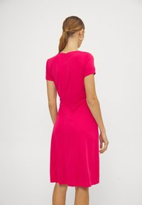 Nainen, jolla on vaaleanruskea hius, sitonut hiuksensa taakse, pukeutuneena kirkkaanpunaiseen lyhythihaiseen polvipituiseen mekkoon, selin valkoista taustaa vasten.