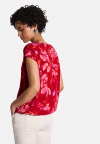 Blusa rossa a maniche corte con motivi floreali rosa, in tessuto leggero, collo rotondo e vestibilità rilassata, abbinata a pantaloni chiari.