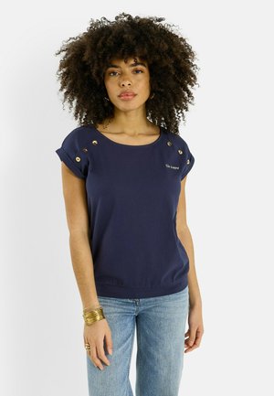 Mujer con cabello rizado que lleva una camiseta de manga corta azul marino con botones dorados y vaqueros azul claro, con accesorios de pulseras y anillo dorados.