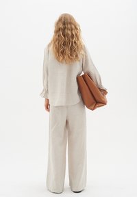 Top in lino color beige chiaro con maniche lunghe e cuciture elastiche, abbinato a pantaloni in lino beige a gamba larga. Borsa a tracolla in pelle marrone tenuta nella mano destra.