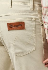 Jeans de mezclilla beige con un parche de cuero con el logo de Wrangler en el bolsillo trasero, que presenta un patrón de costura distintivo por encima del bolsillo.