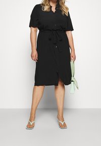 Nainen, jolla on päällä musta napitettu mekko, johon kuuluu vyötärölle sidottu nyöritys. Hänellä on kädessään mintunvihreä käsilaukku ja jalassaan matching mintunvihreät sandaalit.