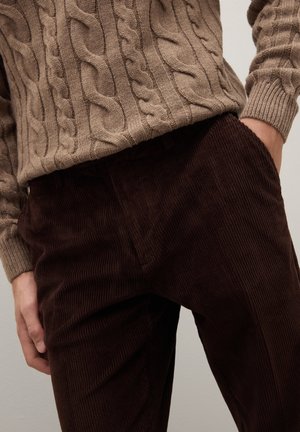 Suéter beige de punto grueso y pantalones de pana marrón oscuro con textura acanalada; los pantalones tienen bolsillos laterales y un ajuste relajado.