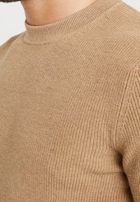 Pull en tricot de couleur marron clair, avec un col rond et une texture côtelée. Design ajusté avec de subtils lignes horizontales.