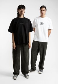 Svart oversized t-skjorte med brodert logo, kombinert med olivengrønne kordfløyelsbukser. Hvit t-skjorte med grafisk design og svarte detaljer.