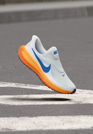 Hvit og blå Nike løpesko med klar oransje såle svever over asfalt med hvite veikryssingsmarkeringer.