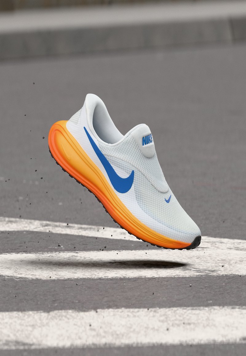 Λευκό και μπλε παπούτσι για τρέξιμο της Nike με φωτεινή πορτοκαλί σόλα να αιωρείται πάνω από άσφαλτο με λευκές οδικές σημάνσεις.