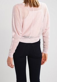 Pull léger rose, en maille lâche, avec un motif texturé, des poignets et une taille côtelés. Porté avec un jean skinny noir, montrant une coupe décontractée.