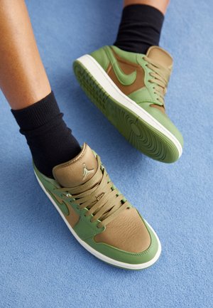 Piernas con calcetines negros y zapatillas Nike verdes y marrones sobre una alfombra azul, un pie levantado mostrando la suela del zapato.