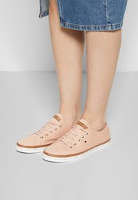Pieds portant des baskets à lacets rose clair avec une bordure marron et un petit logo, associées à une jupe en jean longueur genou sur un sol uni.
