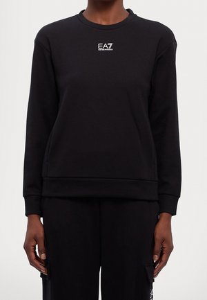 Personne portant un sweat-shirt à manches longues noir et un pantalon assorti, avec un petit logo blanc "EA7 Emporio Armani" sur la poitrine du sweat-shirt.