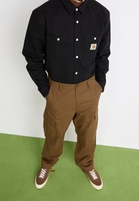 Schwarzes Hemd mit Knöpfen und zwei Brusttaschen; braune Cargo-Hosen mit Seitentaschen; braune Sneakers mit weißen Schnürsenkeln; grüner Boden als Hintergrund.