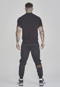 Zwarte crew-neck t-shirt met merkgele tekst op de mouwen, gecombineerd met zwarte joggers. Heeft tatoeages en zwart-witte sneakers.