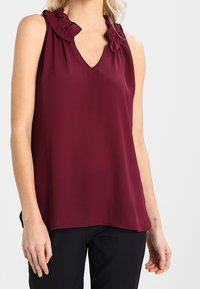 Top senza maniche di colore bordeaux con scollatura a V e dettagli arricciati lungo le spalle. Realizzato in un tessuto leggero e morbido. Abbinato a pantaloni neri.
