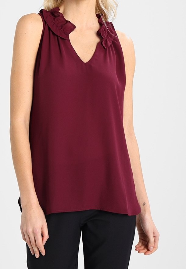 Top senza maniche di colore bordeaux con scollatura a V e dettagli arricciati lungo le spalle. Realizzato in un tessuto leggero e morbido. Abbinato a pantaloni neri.