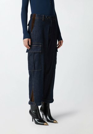Pantaloni cargo in denim blu scuro con tasche laterali, caratterizzati da pannelli laterali marroni, risvolti alle caviglie e dettagli dorati su tacchi neri a punta.