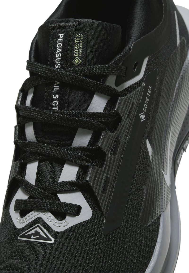 Zapatilla deportiva negra con marca Gore-Tex, que presenta un tejido texturizado, diseño con cordones y acentos reflectantes en la parte superior y zona de los dedos.