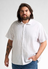 Witte linnen shirt met korte mouwen en een button-down kraag, gladde textuur en een relaxed fit, gecombineerd met blauwe jeans. Bevat zichtbare tatoeages op de arm.